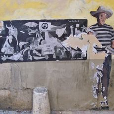 Póster pegado a la pared con una imagen de Pablo Picasso de pie, con sombrero y dos botes de espray junto a su obra 'Guernica'. En el cuadro aparece un símbolo de la paz y el lema «Stop the massacre».