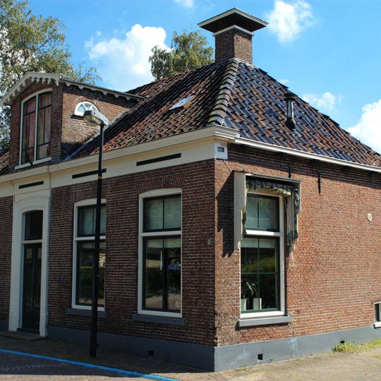 Laag dorpshuis met omlijste ingang, zesruitsvensters en versierde kroonlijst onder schilddak met twee hoekschoorstenen waarop borden