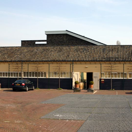 Langgerekte, gesloten loods opgetrokken in hout delen onder pannen zadeldak tussen puntgevels. Te linkerzijde aangekapt een aanbouw onder lessenaarsdak