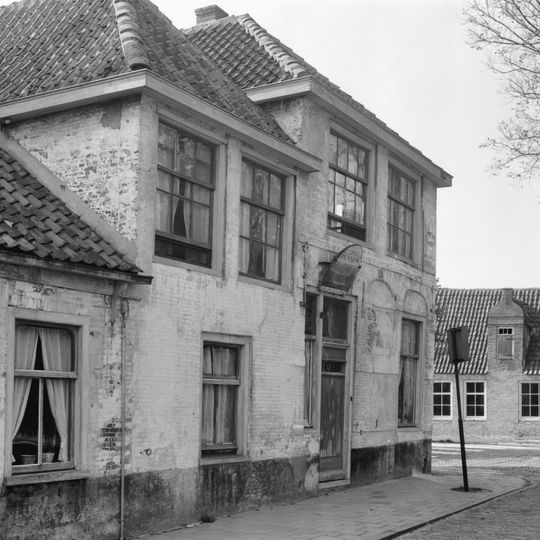 Hoekhuis thans met schilddak. Gevel met vensterbogen en gevelsteen