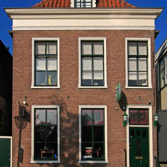 Pand met eenvoudige lijstgevel ter breedte van drie vensterassen. Geprofileerde houten vensteromlijstingen. Stoep met hardstenen palen aan weerskanten