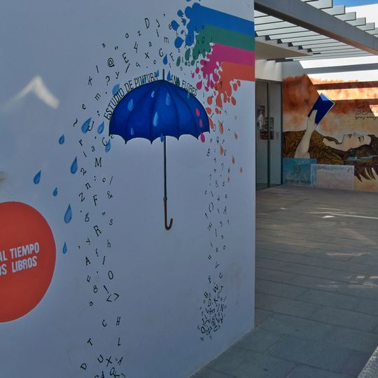 Mural que representa una mujer con los ojos cerrados con un libro abierto entre sus manos y un flujo de agua multicolor que se convierte en una lluvia de letras sobre un paraguas abierto que funciona como fotocol.