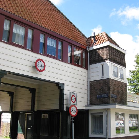Poortgebouw met twee winkels met bovenwoningen, met veel hout zoals de zware kolommen in poort, bouwstijl Amsterdamse School, ligging aan Purmerweg centraal deel van Tuindorp Nieuwendam