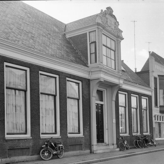 Zeven traveeën breed pand onder schilddak met boven de ingang overgebouwde erker bekroond met snijwerk